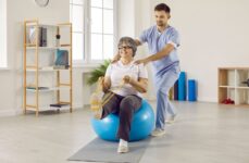 Male physio therapy helping a senior woman with a leg/core exercise, using a band and ball.