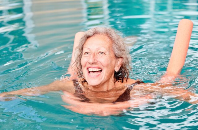 An older adult smiling and swimming in the community pool with the assistance of an orange pool noodle.